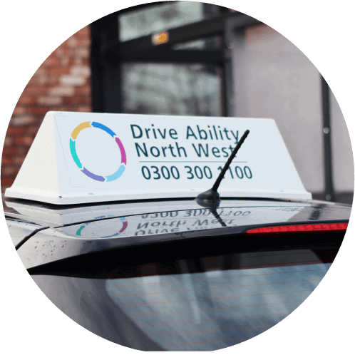 A car roof sign reads “Drive Ability North West” with a contact number and a colorful circular logo. The vehicle is parked near a brick building with a glass door.
