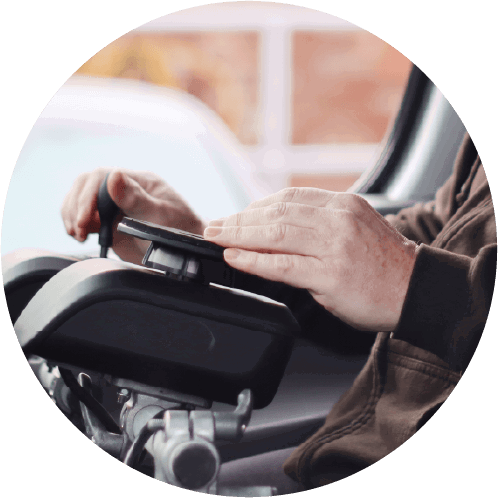 A person in a brown jacket uses a joystick control panel, likely for a powered wheelchair, with a blurred indoor background and window view.
