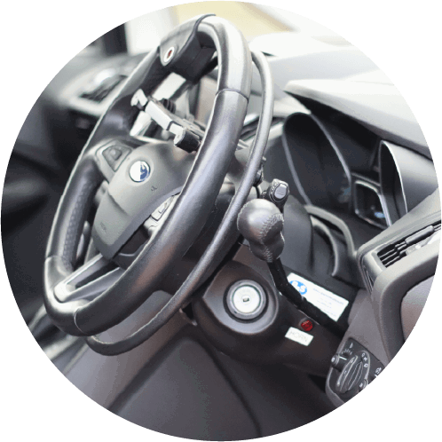 Close-up of a car’s steering wheel fitted with a spinner knob and hand control lever, designed for drivers with physical disabilities. The dashboard, ignition, and control buttons are visible.